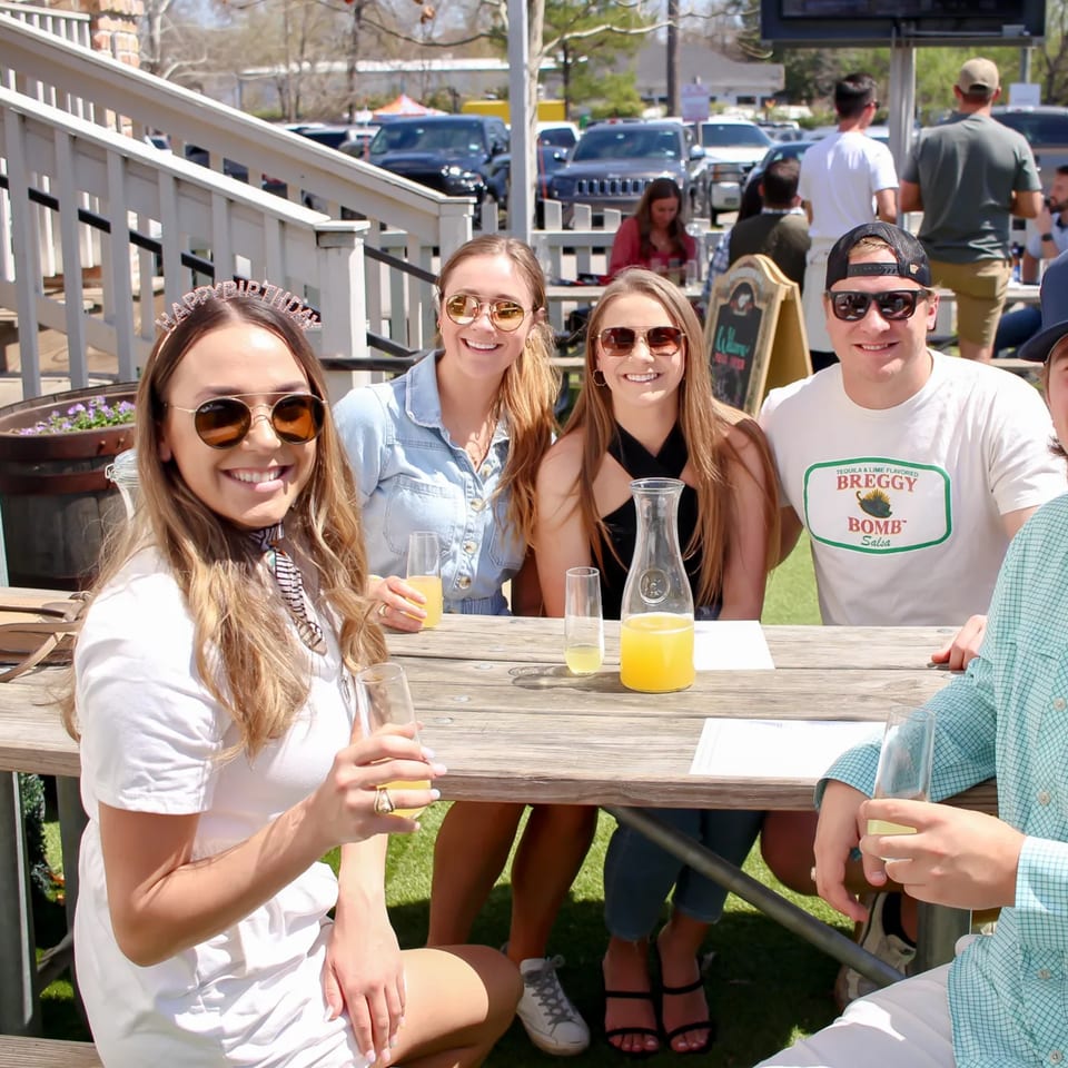 Four young adults enjoying mimosas at an outdoor picnic table on a sunny day at Bungalow Heights.