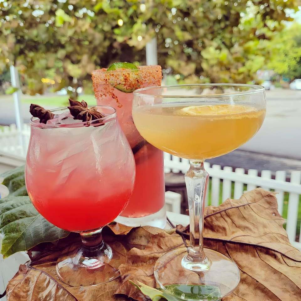 Three colorful cocktails, two pink and one yellow, on a table with large leaves and a white fence.