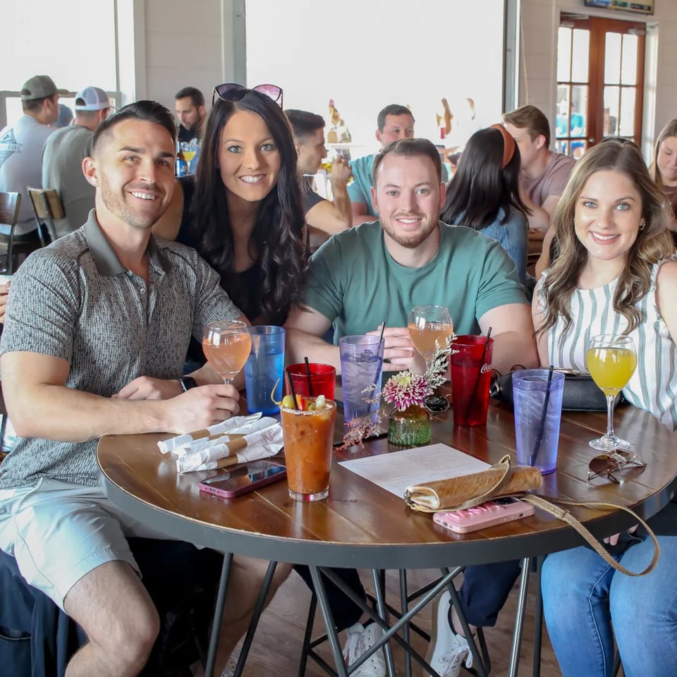 Four smiling friends enjoying drinks at a round table in a lively bar-and-grill setting.