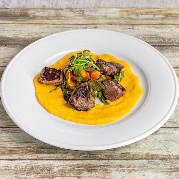 Grilled steak pieces on a bed of yellow puree, topped with a fresh green and orange salad.