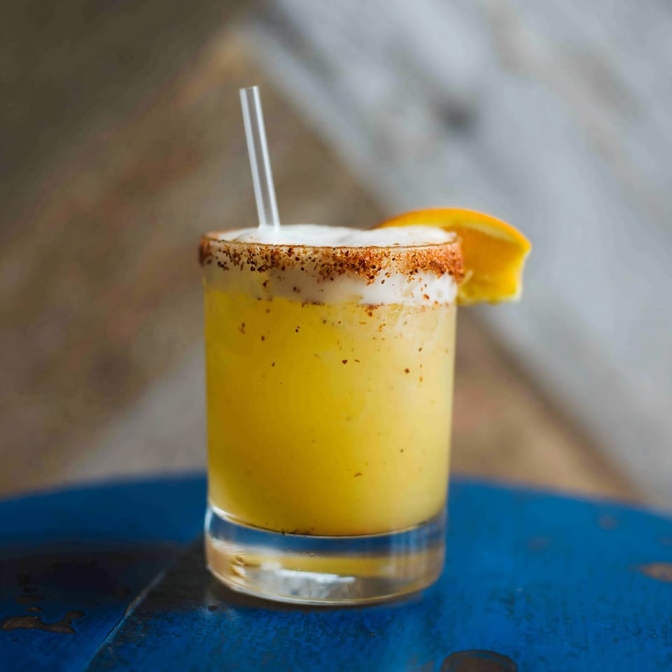 A Bungalow Heights cocktail, yellow with a spiced rim, orange slice, and straw on a blue table.
