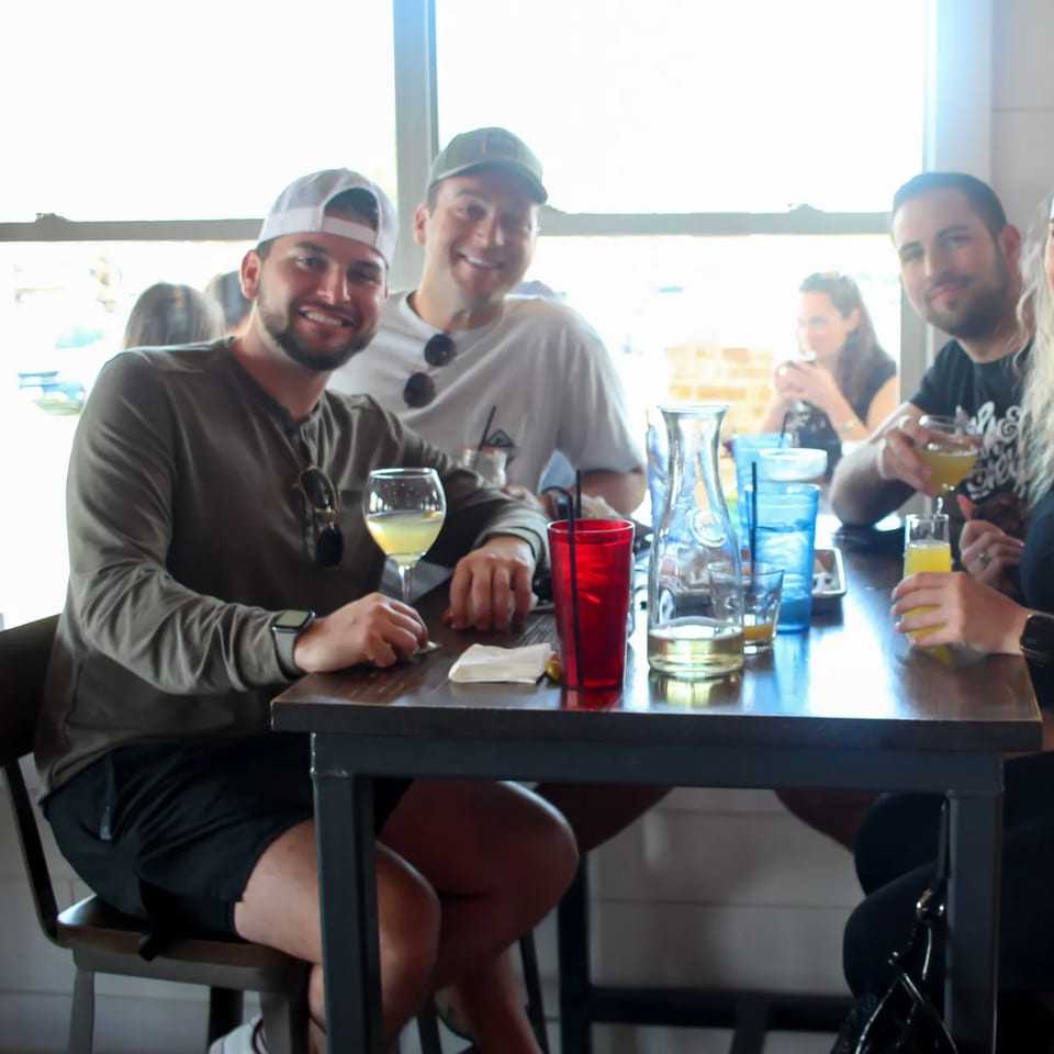 Four smiling people enjoying drinks at a table in a bright, casual bar-and-grill setting.