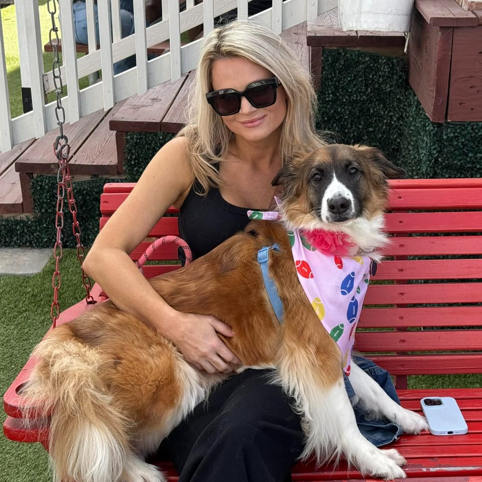A woman in sunglasses sits on a red swing with a brown and white dog wearing a bandana.