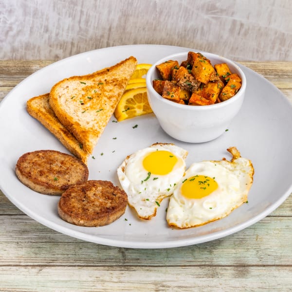 A breakfast plate with two fried eggs, two sausage patties, toast, orange slices, and roasted potatoes.
