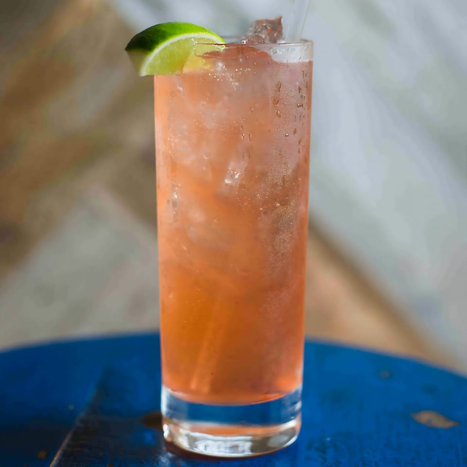 A tall glass of iced pink cocktail with a lime wedge on a blue table at Bungalow Heights.