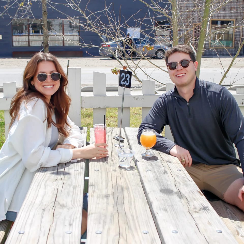 Two smiling people with drinks at a wooden picnic table on the Bungalow Heights patio.