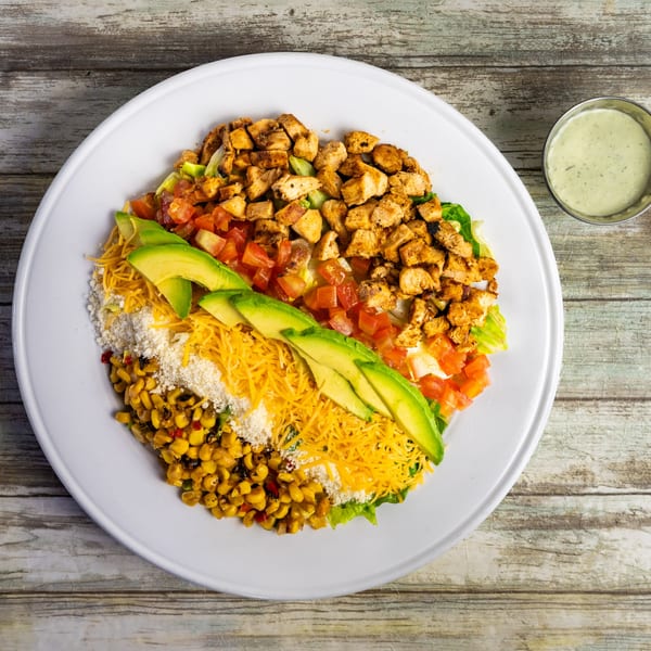 A vibrant Bungalow Heights salad with grilled chicken, corn, avocado, cheese, and tomatoes, with dressing on the side.