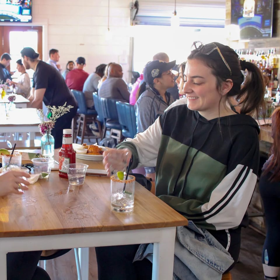 A woman smiles at a table with drinks and food, surrounded by other patrons at Bungalow Heights.