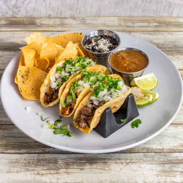 Three tacos with meat, onions, and cilantro, served with chips, black beans, salsa, and lime wedges.