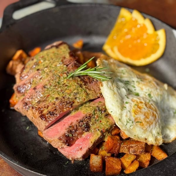 Sliced steak with green sauce, fried eggs, roasted potatoes, and an orange slice in a skillet.