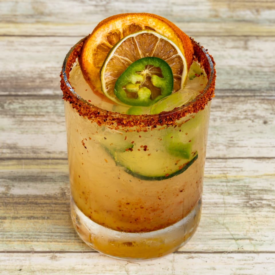 Spicy cocktail with a chili-lime rim, garnished with dried orange, lime, and fresh jalapeño slices.