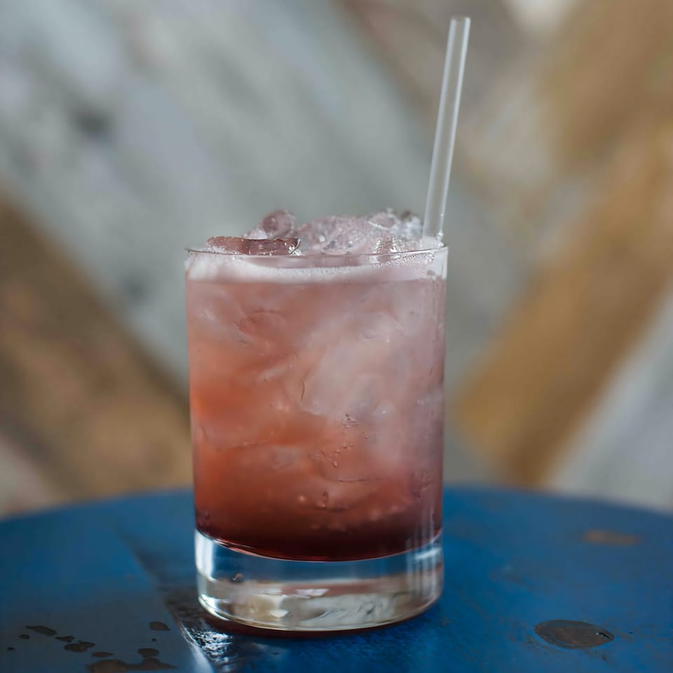 A vibrant red cocktail with ice and a clear straw sits on a blue table at Bungalow Heights.